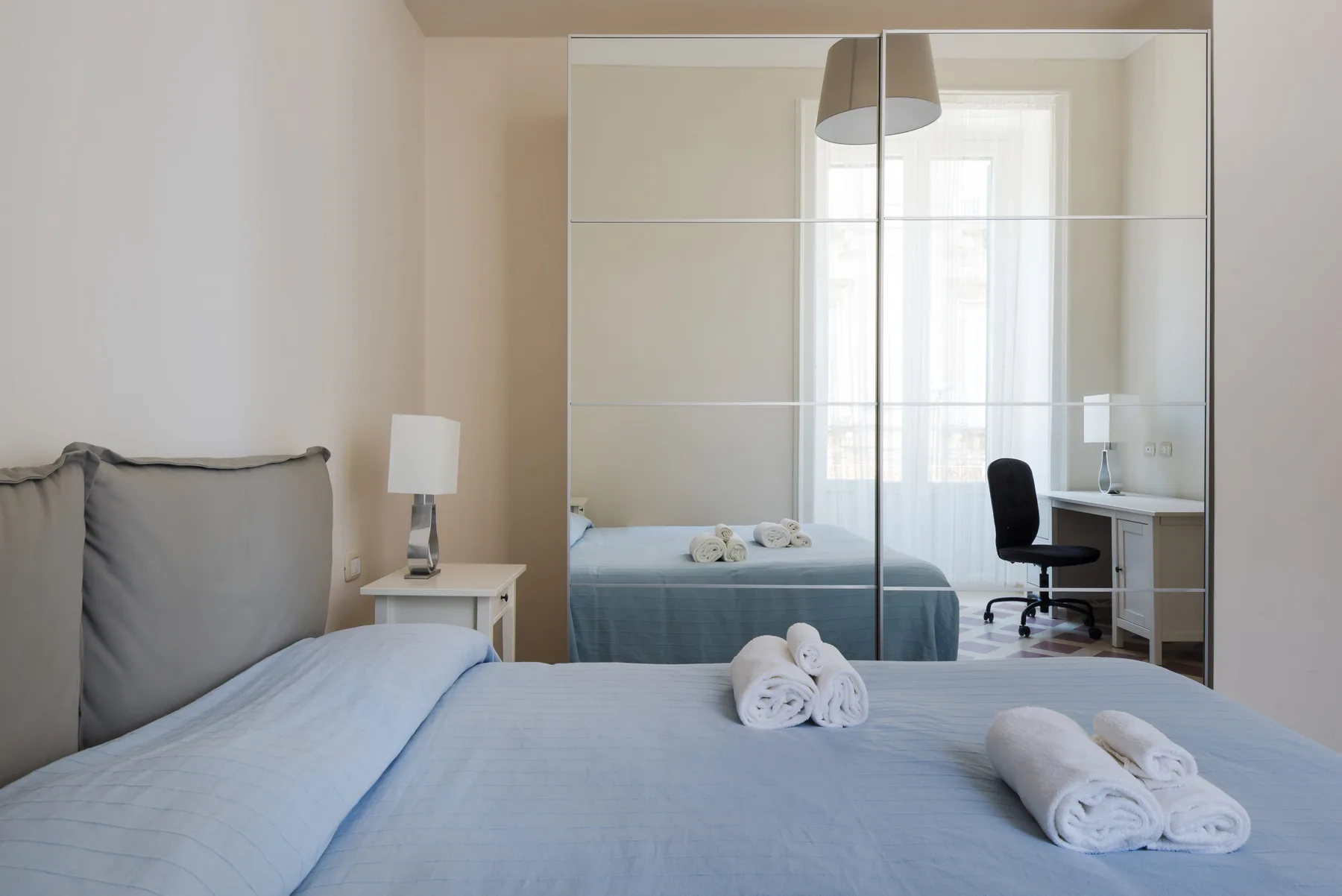 Un dormitorio luminoso con una cama, un armario con espejo y un escritorio junto a la ventana.