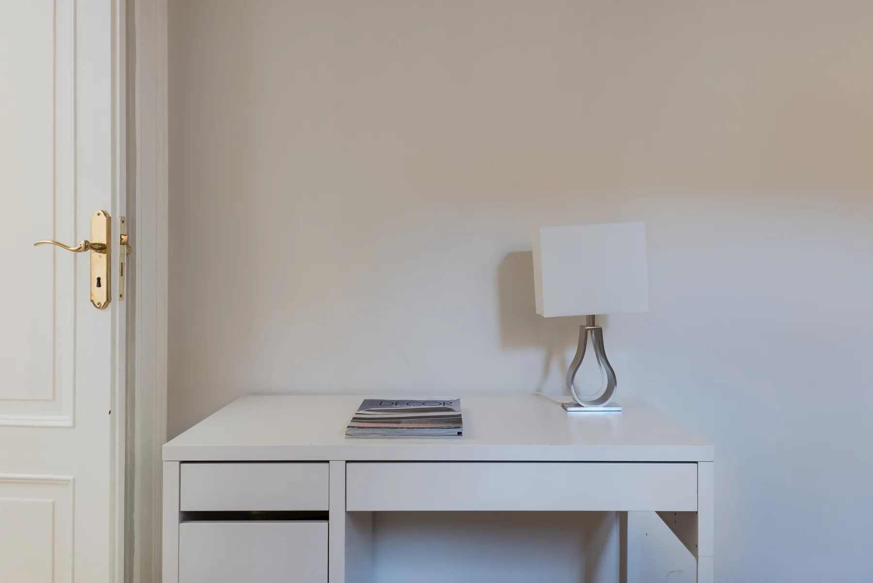 Un escritorio blanco con una lámpara de mesa y libros apilados está junto a una puerta con paneles.