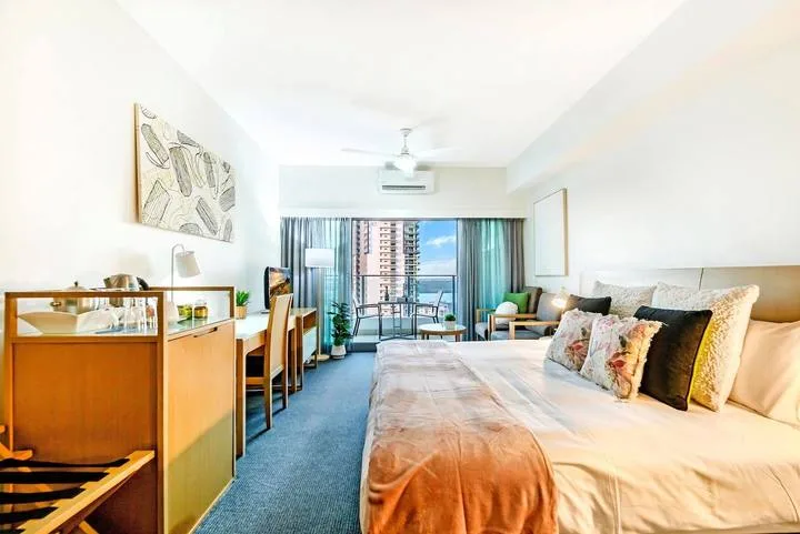 A bright bedroom with a large bed, desk, and balcony doors overlooking the city.