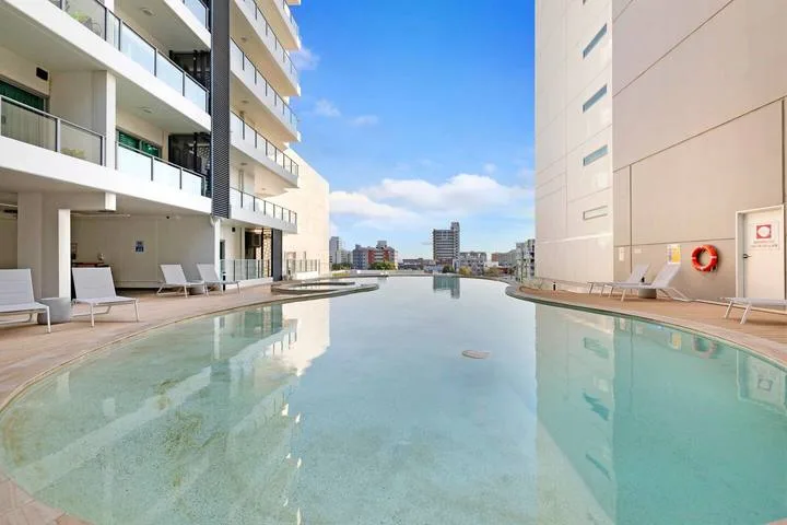 An outdoor swimming pool between modern buildings, with loungers along the edge.