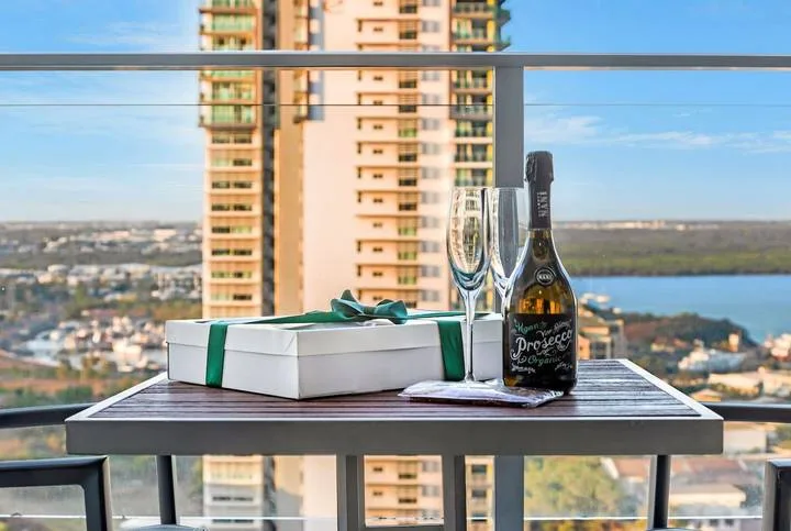 A balcony table displays a gift box, champagne bottle, and glasses against a city view.
