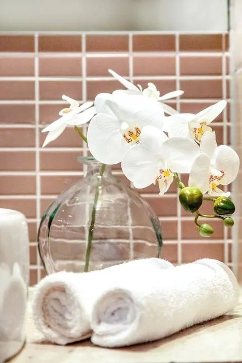 White orchids in a glass vase sit behind rolled towels against a tiled wall.