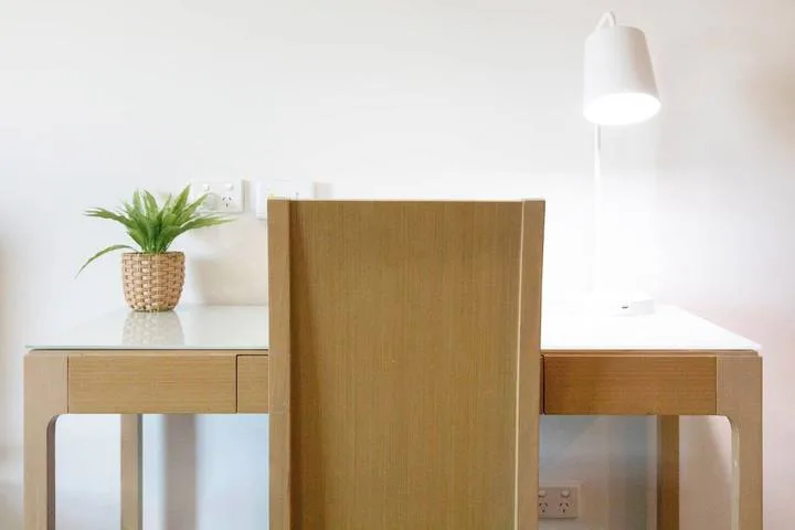 A wooden desk and chair sit beneath a wall lamp, with a small potted plant nearby.