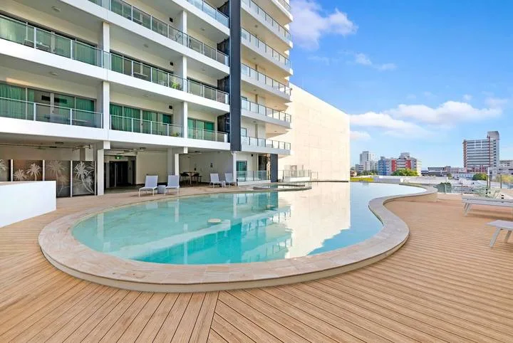 A swimming pool curves beside a multi-story building and a wooden deck.