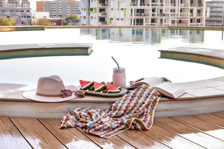 A poolside deck with a hat, a drink, watermelon, and a draped towel.
