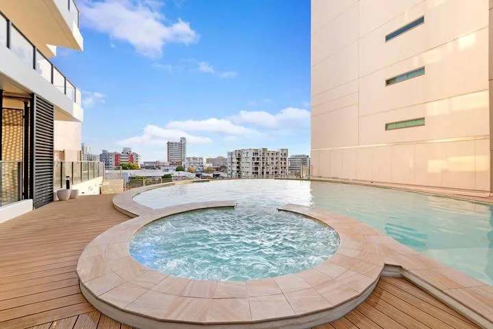 A rooftop pool with a circular spa and wooden decking overlooks nearby buildings.