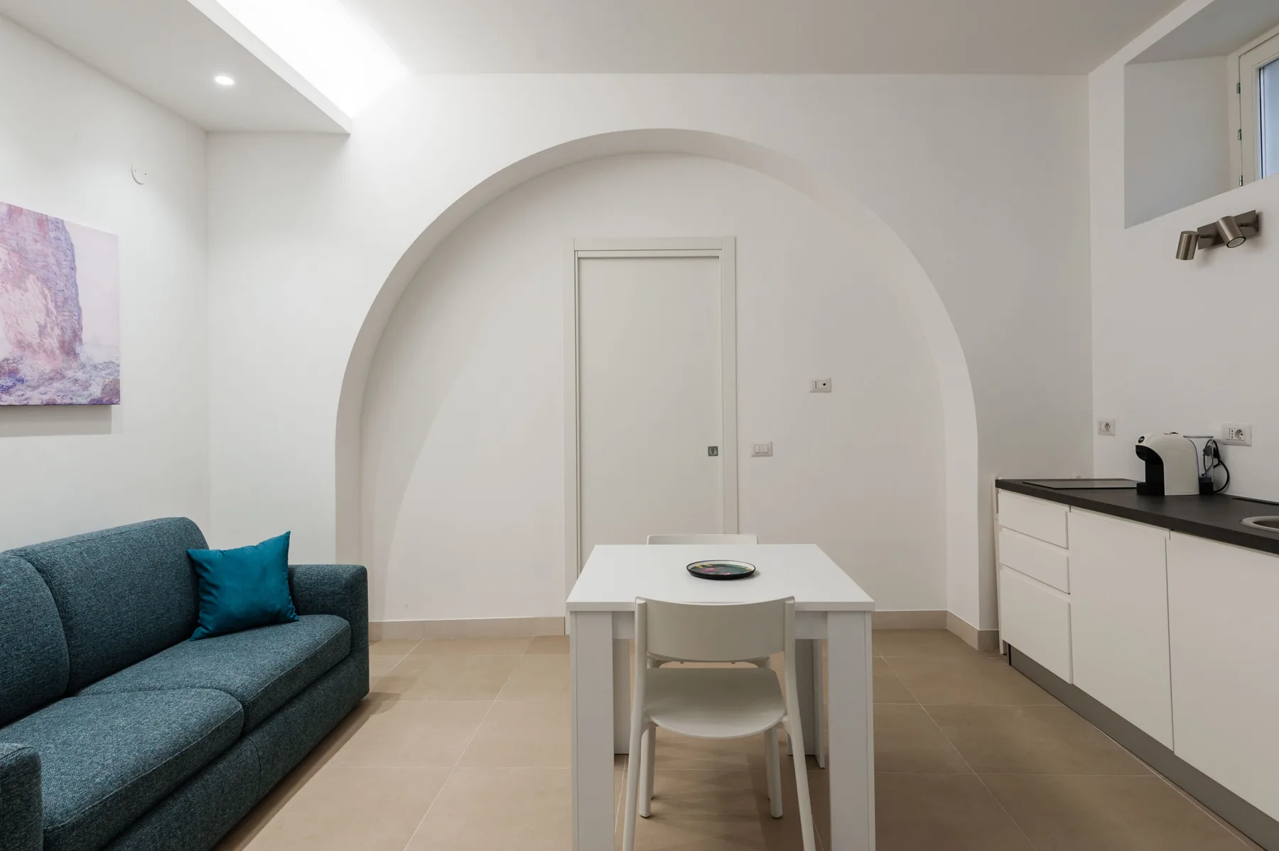 Una sala luminosa tiene un sofá verde azulado, una mesa pequeña y una encimera de cocina.