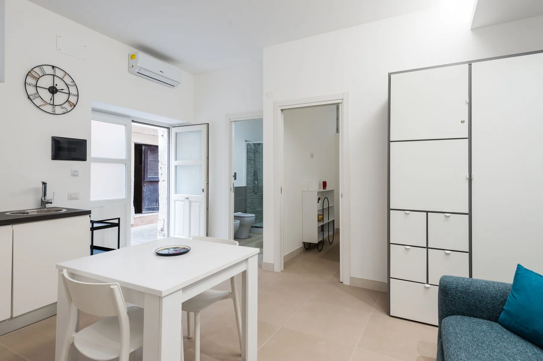 Un comedor luminoso y blanco, con una mesa y asientos junto a una puerta abierta.