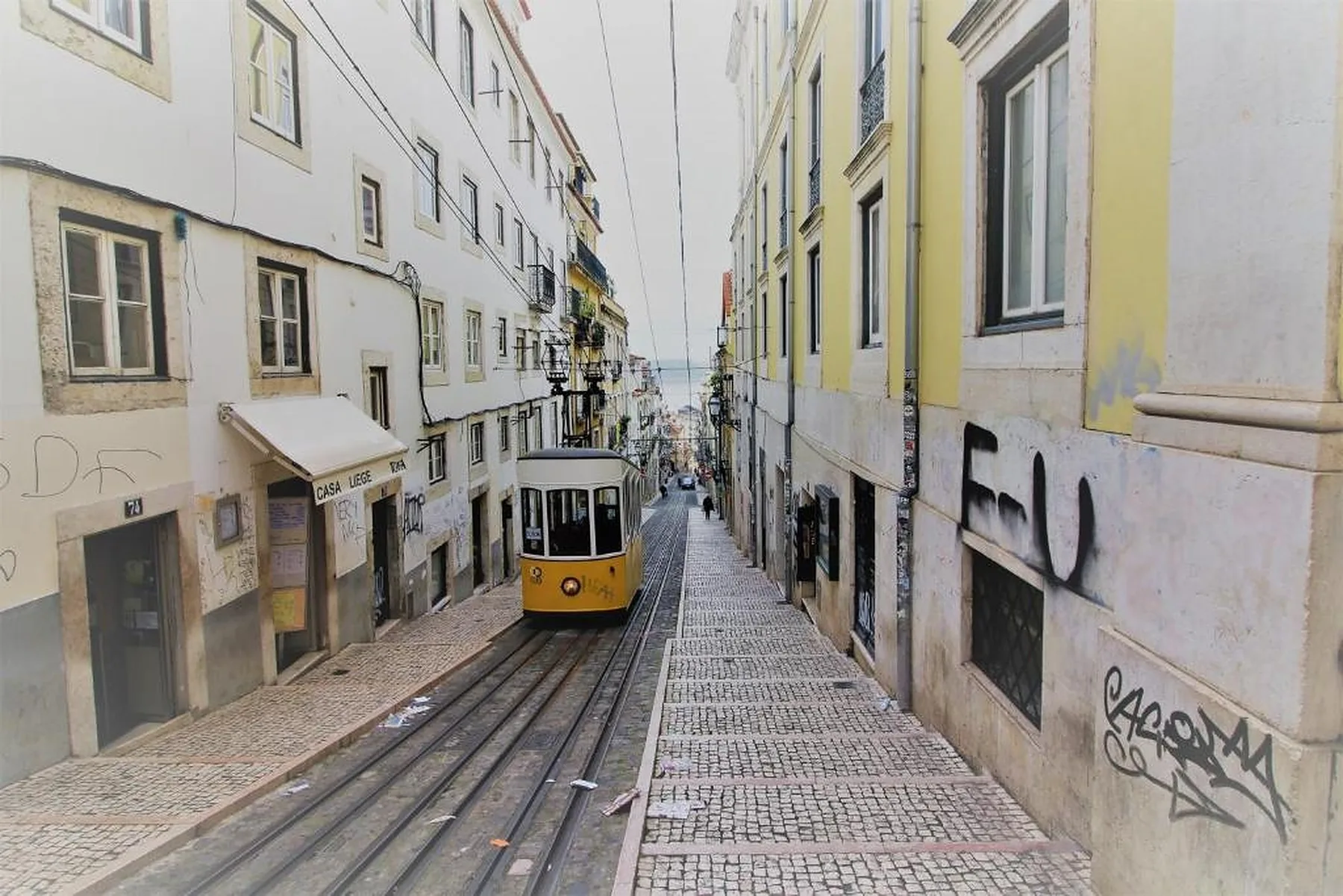 A narrow Lisbon street slopes downhill between buildings, with a yellow tram on the tracks.