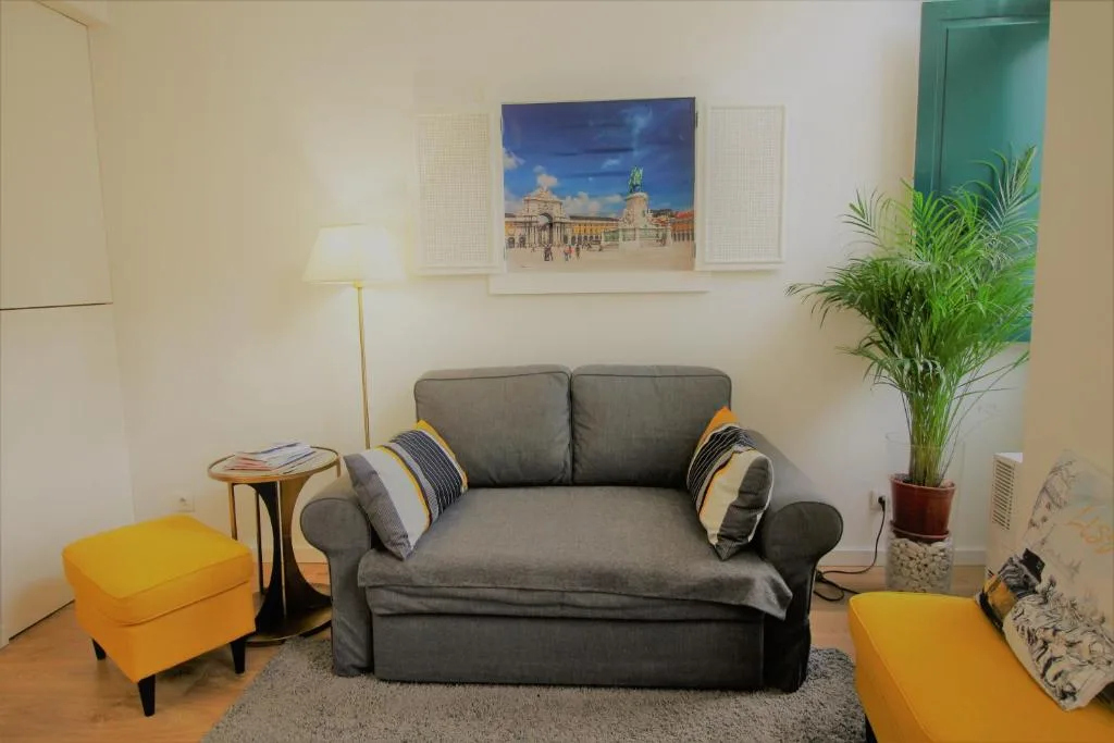 Una sala de estar con un sofá gris, un reposapiés amarillo y una planta en maceta.