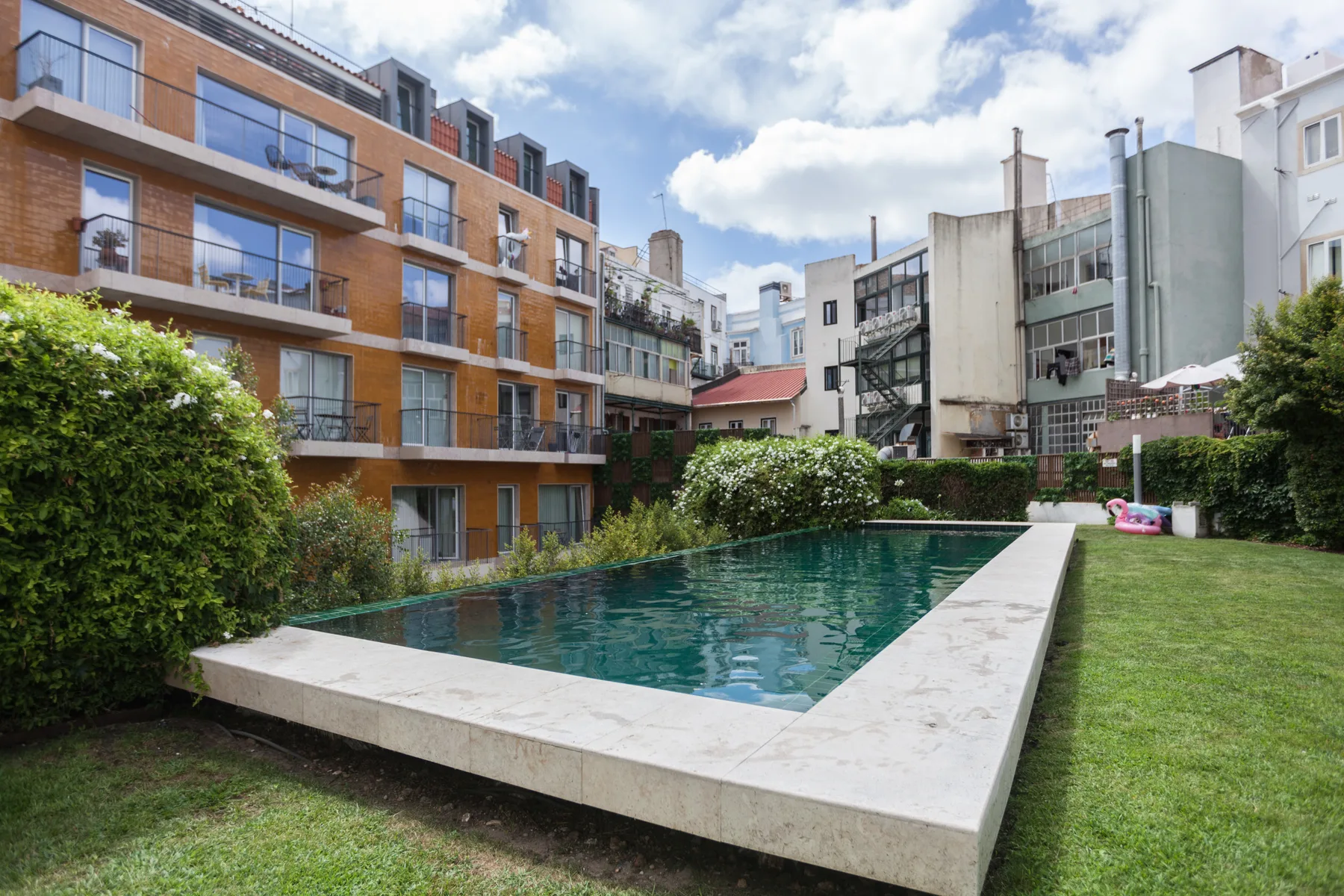 Una piscina al aire libre está junto a un césped y edificios de apartamentos.