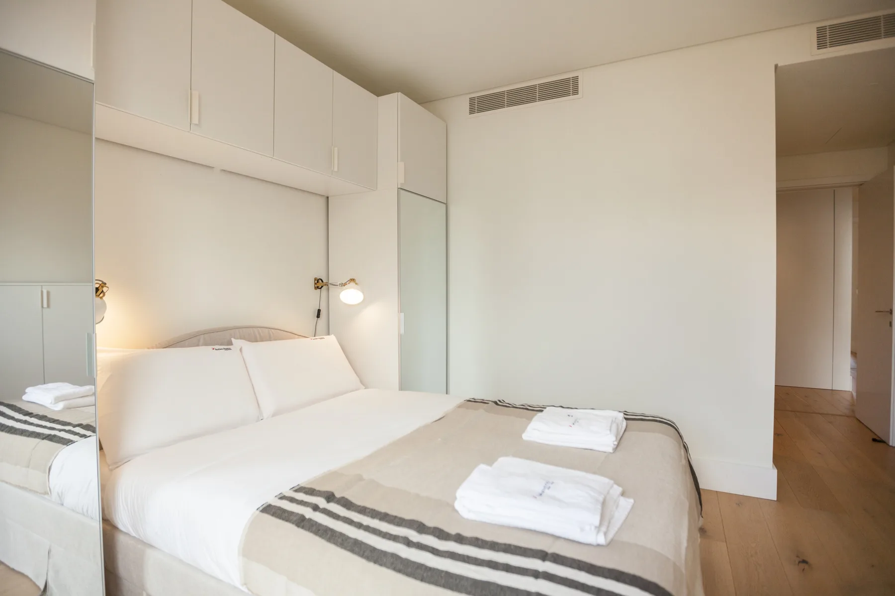 Dormitorio con una cama bien hecha, toallas dobladas y una lámpara de lectura en la pared.