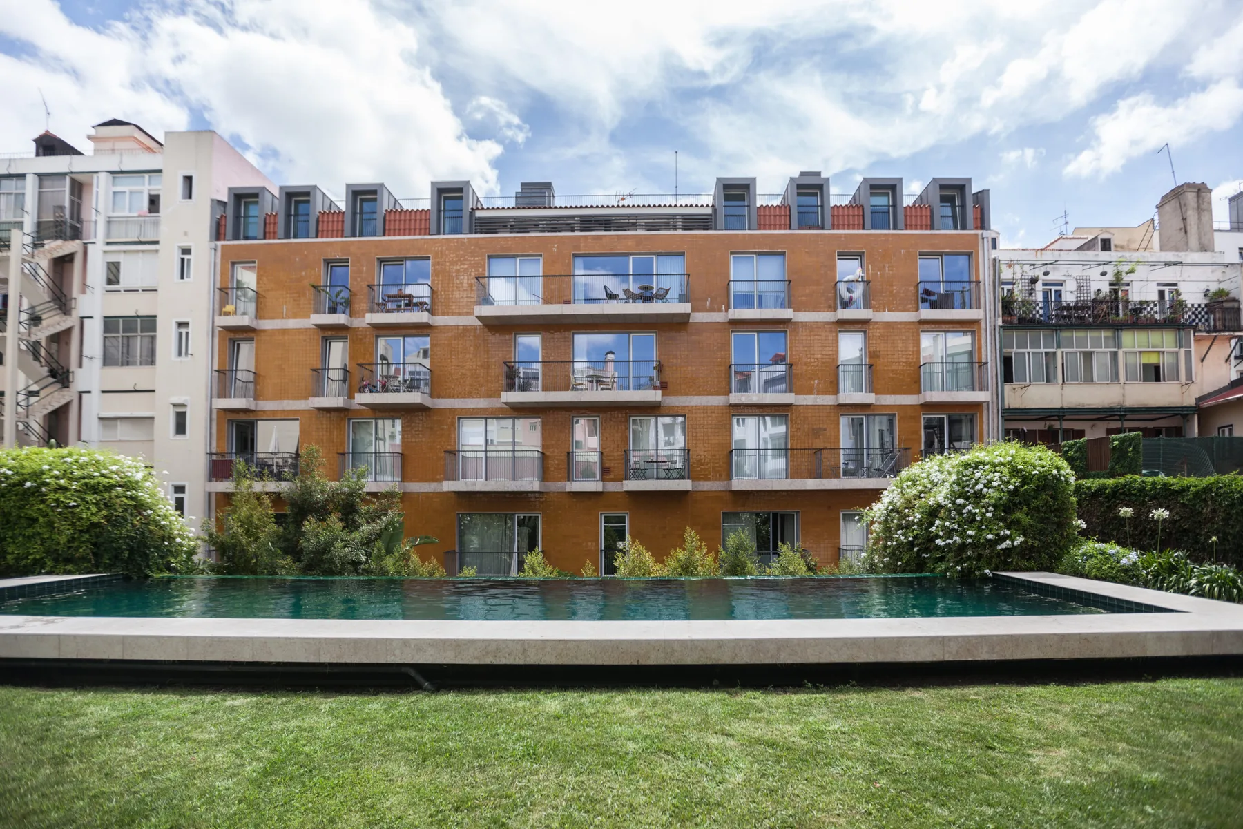 Un edificio residencial se alza detrás de una piscina y un patio con césped.