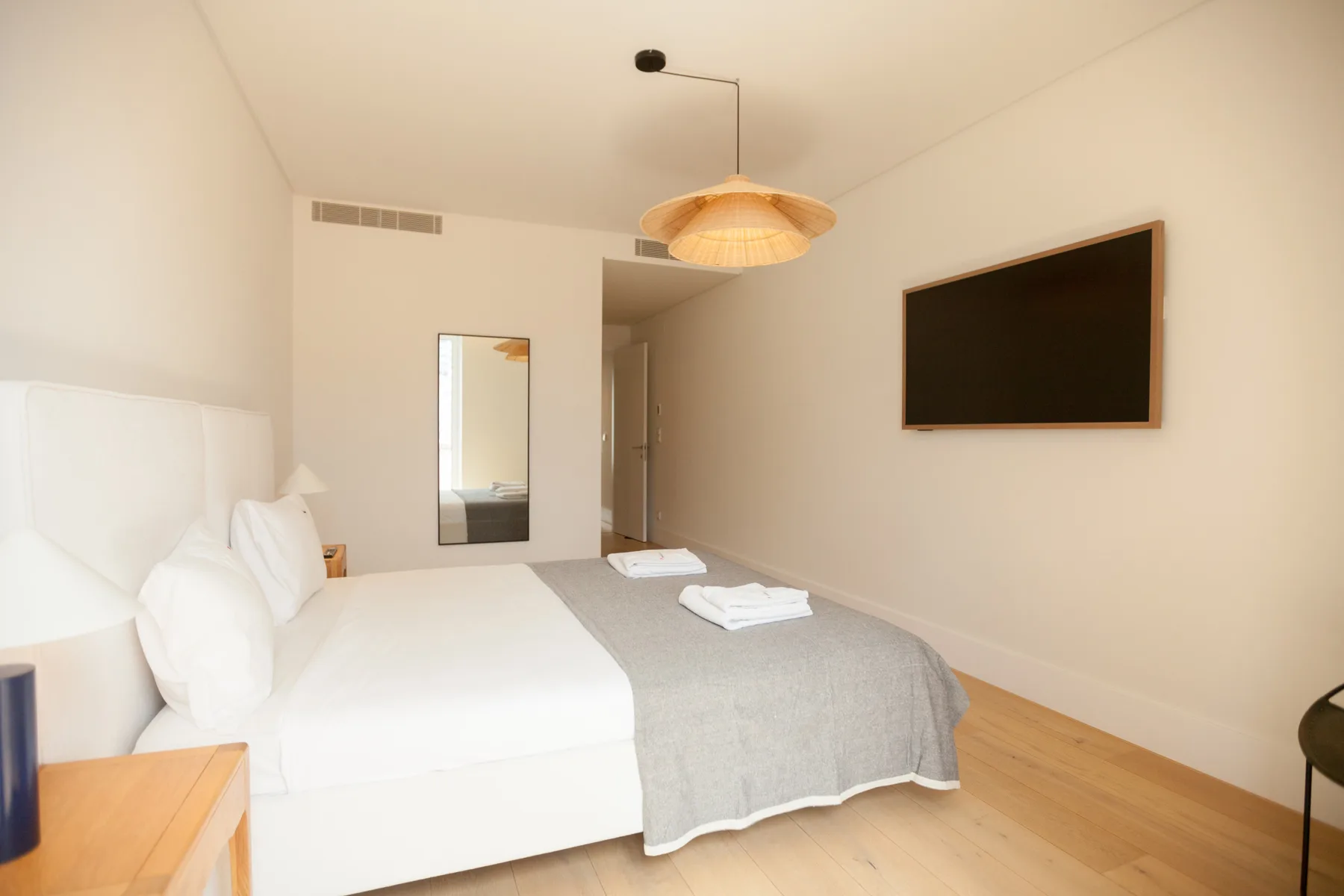 Un dormitorio luminoso con una cama, una colcha gris y un televisor en la pared.
