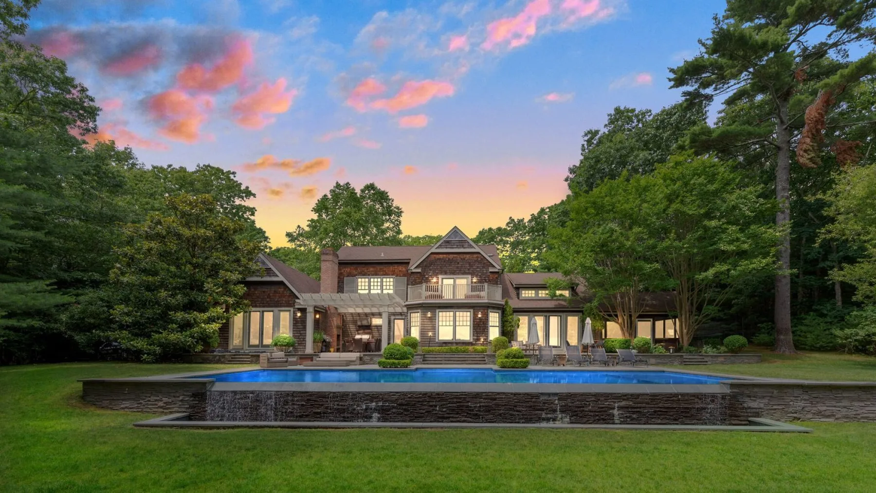 Una casa grande se alza detrás de una piscina rectangular al atardecer.