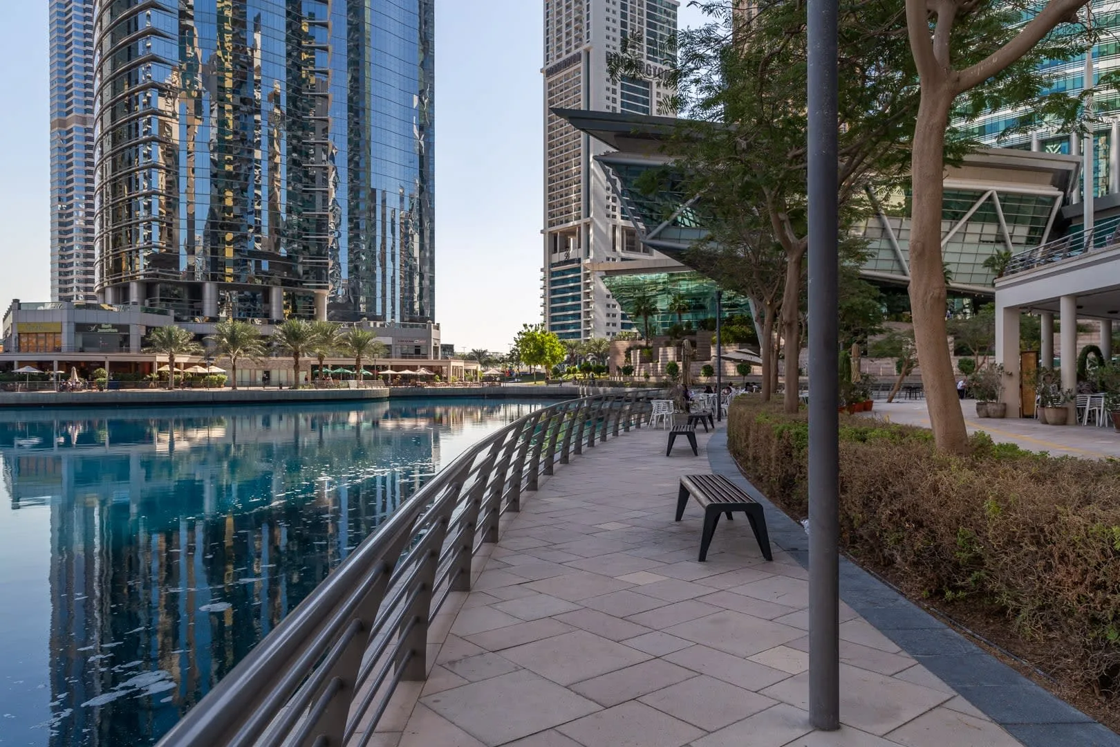 Un paseo pavimentado con bancos bordea una piscina reflectante junto a edificios modernos.