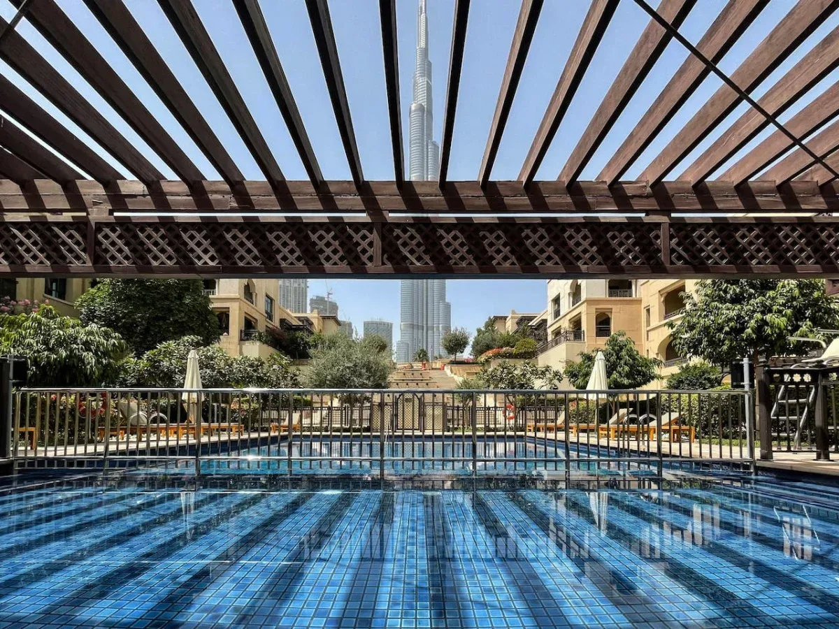 A swimming pool beneath a slatted pergola, with a tall tower visible in the distance.