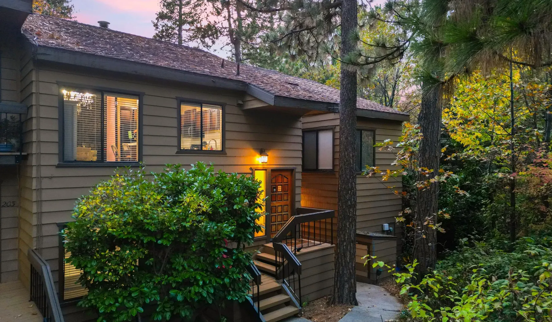 A wood-sided cabin exterior with lit windows, a small entry deck, and surrounding trees.