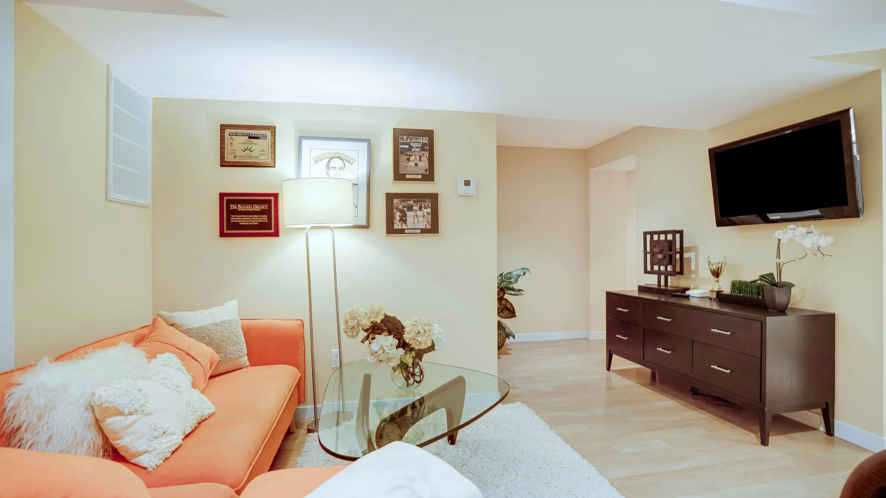 Una sala de estar con asientos naranjas, una mesa de centro de vidrio y un televisor en la pared.