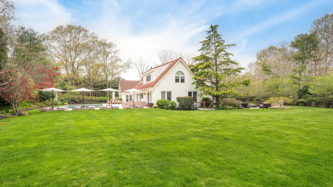 Una casa blanca se alza tras un amplio césped verde, con árboles y asientos de patio cerca.
