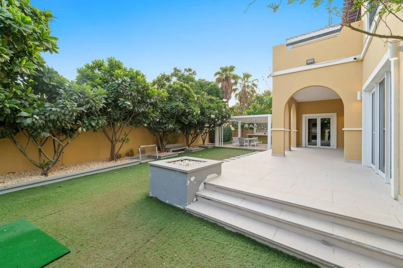 Un patio embaldosado junto al césped, con una entrada en arco y árboles frondosos.