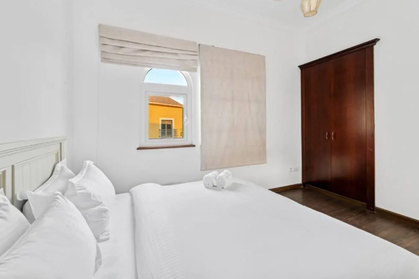 Un dormitorio luminoso con una cama blanca y un armario de madera oscura.