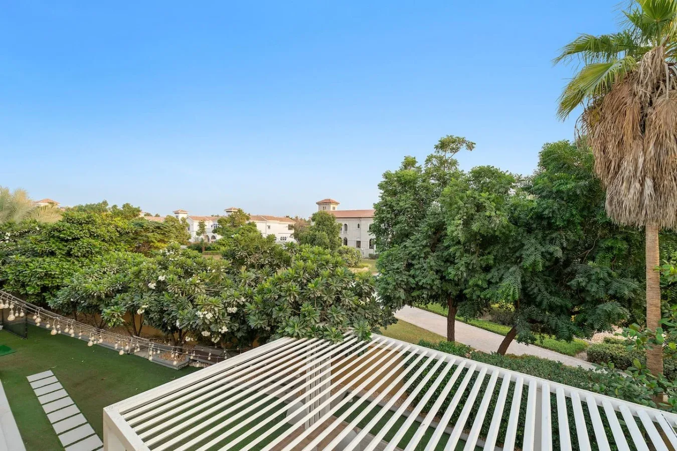 Un jardín con árboles frondosos, una palmera y una pérgola blanca bajo un cielo despejado.