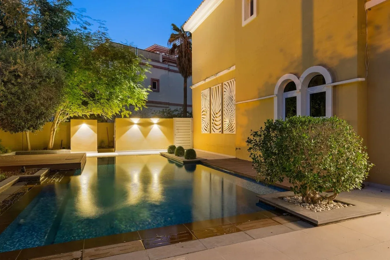 Una piscina junto a un edificio amarillo, rodeada de árboles y arbustos.