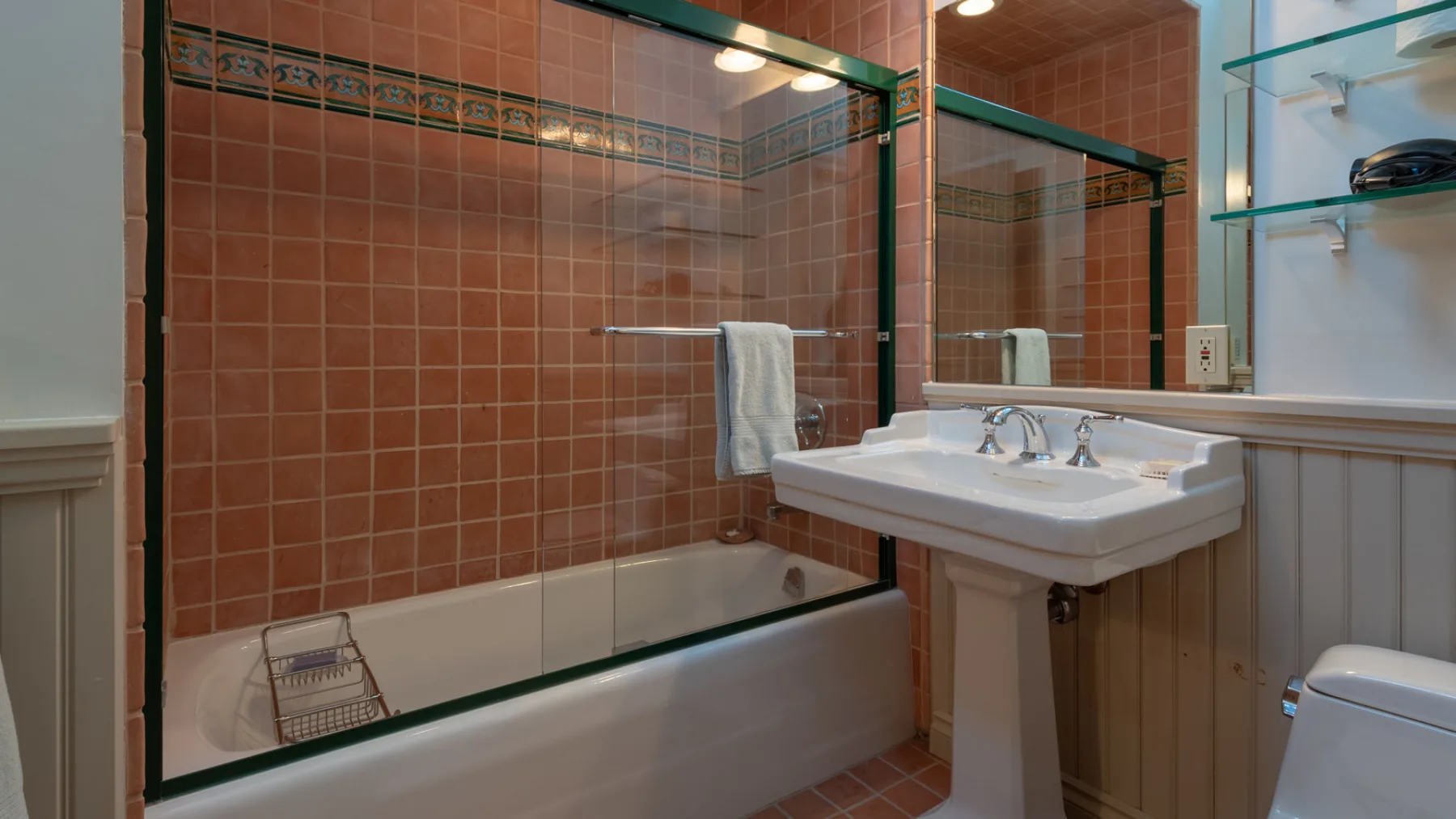 A bathroom with a tiled bathtub, pedestal sink, and mirror.