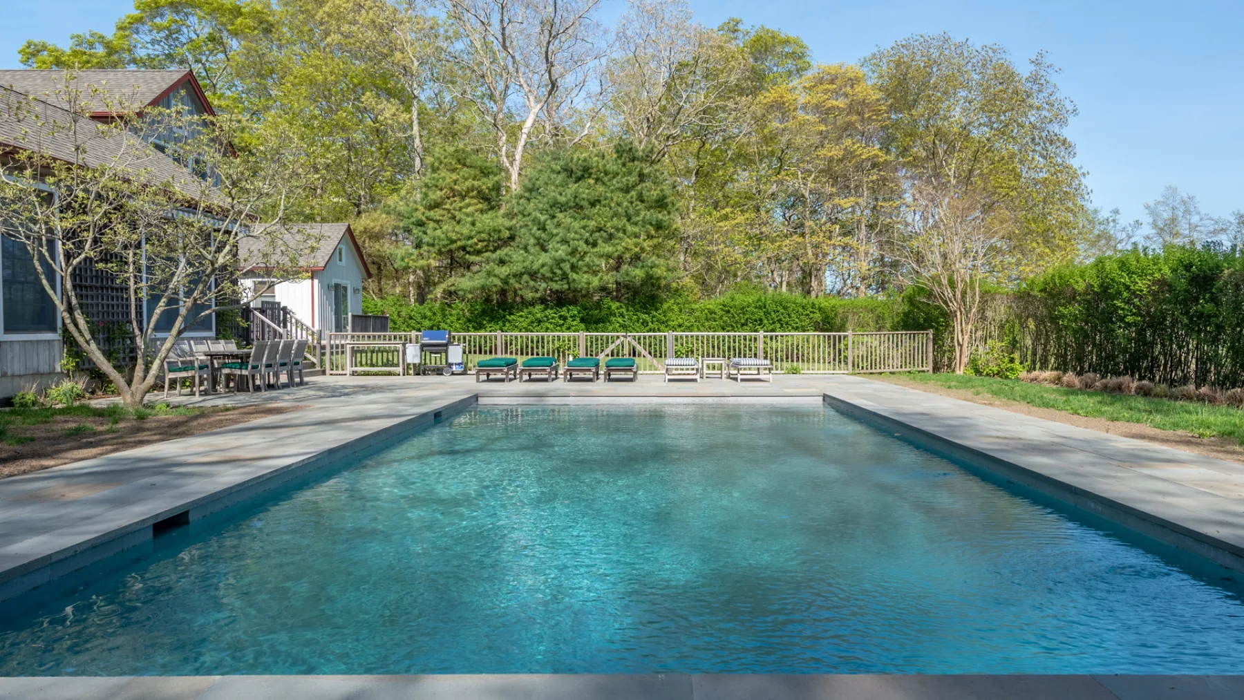 An outdoor swimming pool is surrounded by a paved deck, seating, and trees.