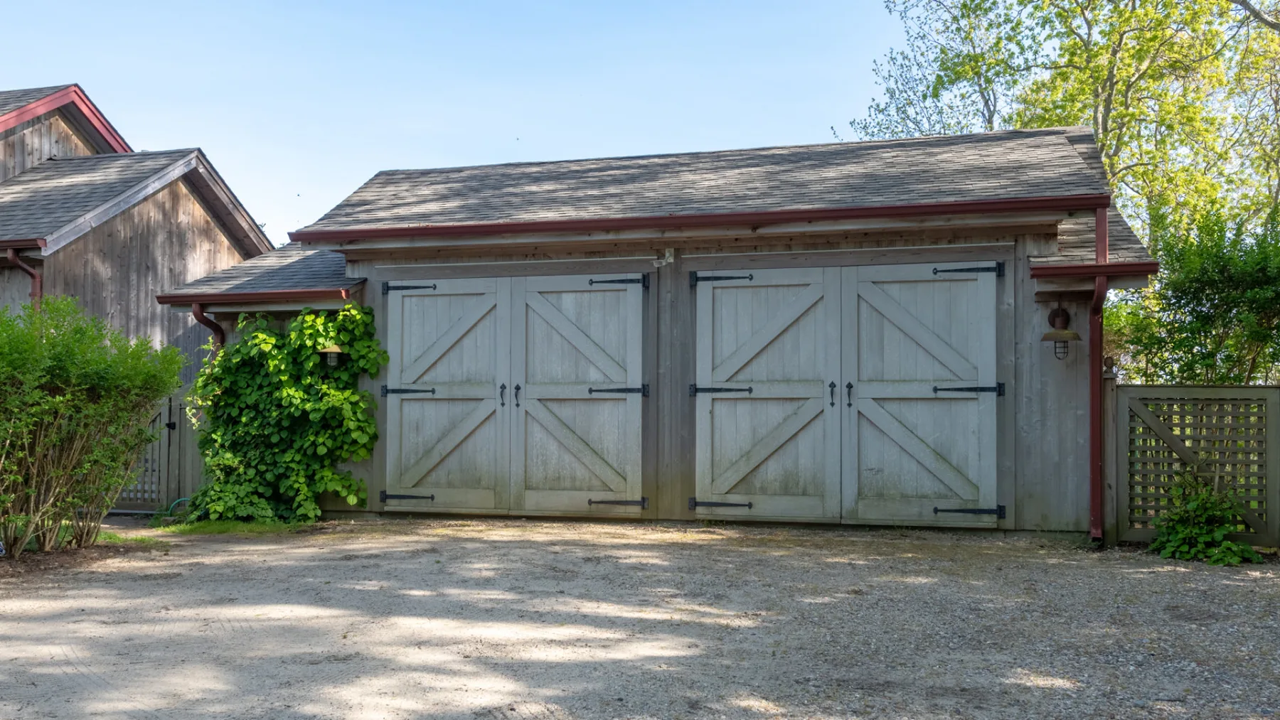 A gray wooden outbuilding with double doors stands beside a gravel driveway.