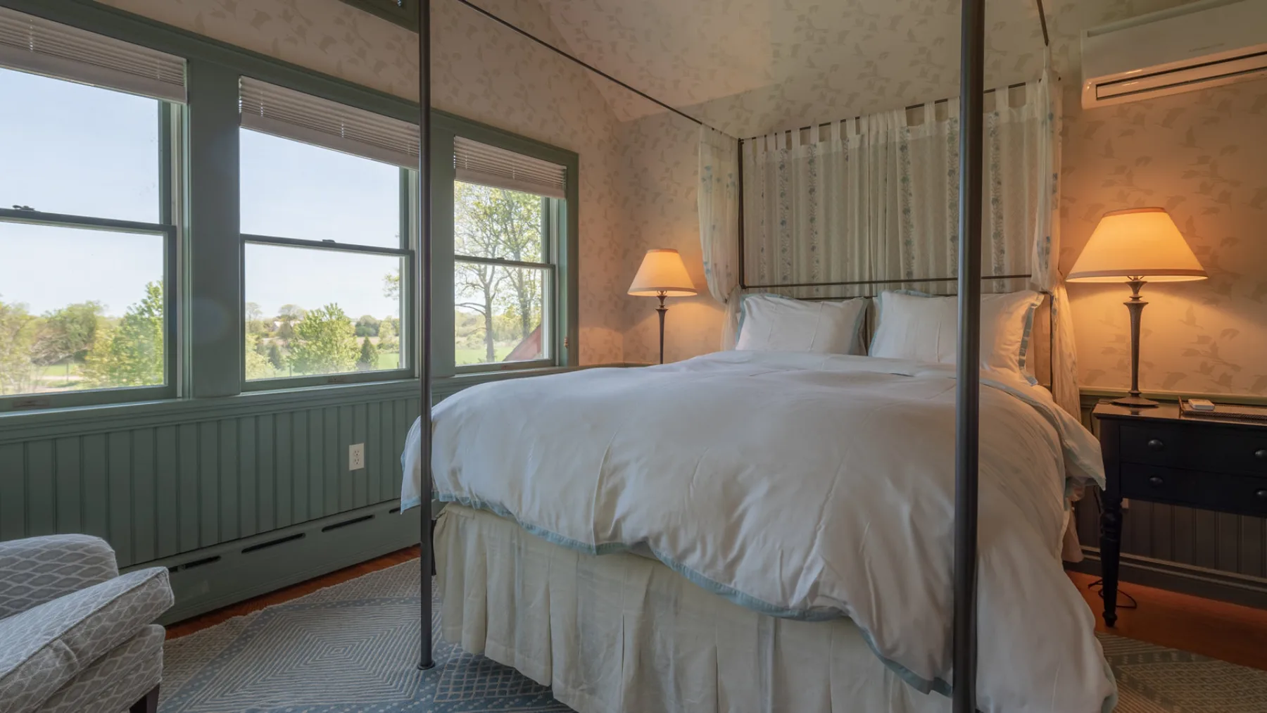 A bedroom with a large bed, bedside lamps, and windows overlooking trees.