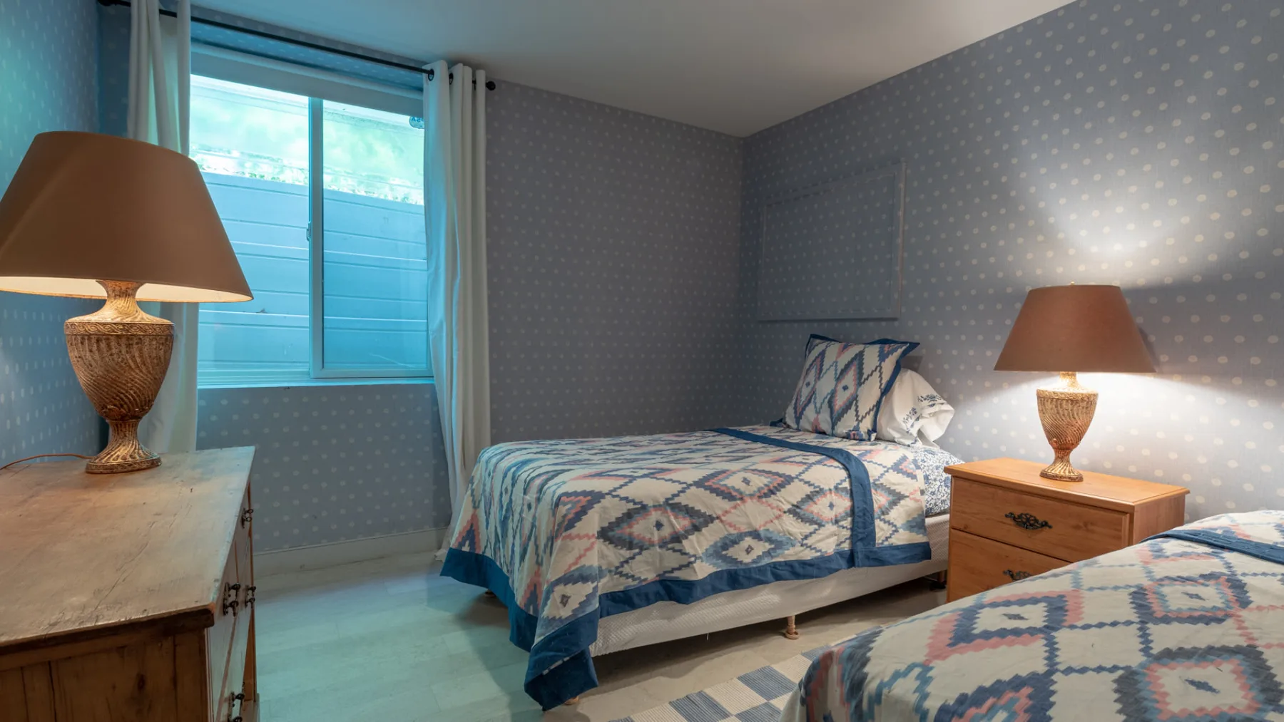 A bedroom with a bed dressed in a blue patterned quilt and matching curtains.