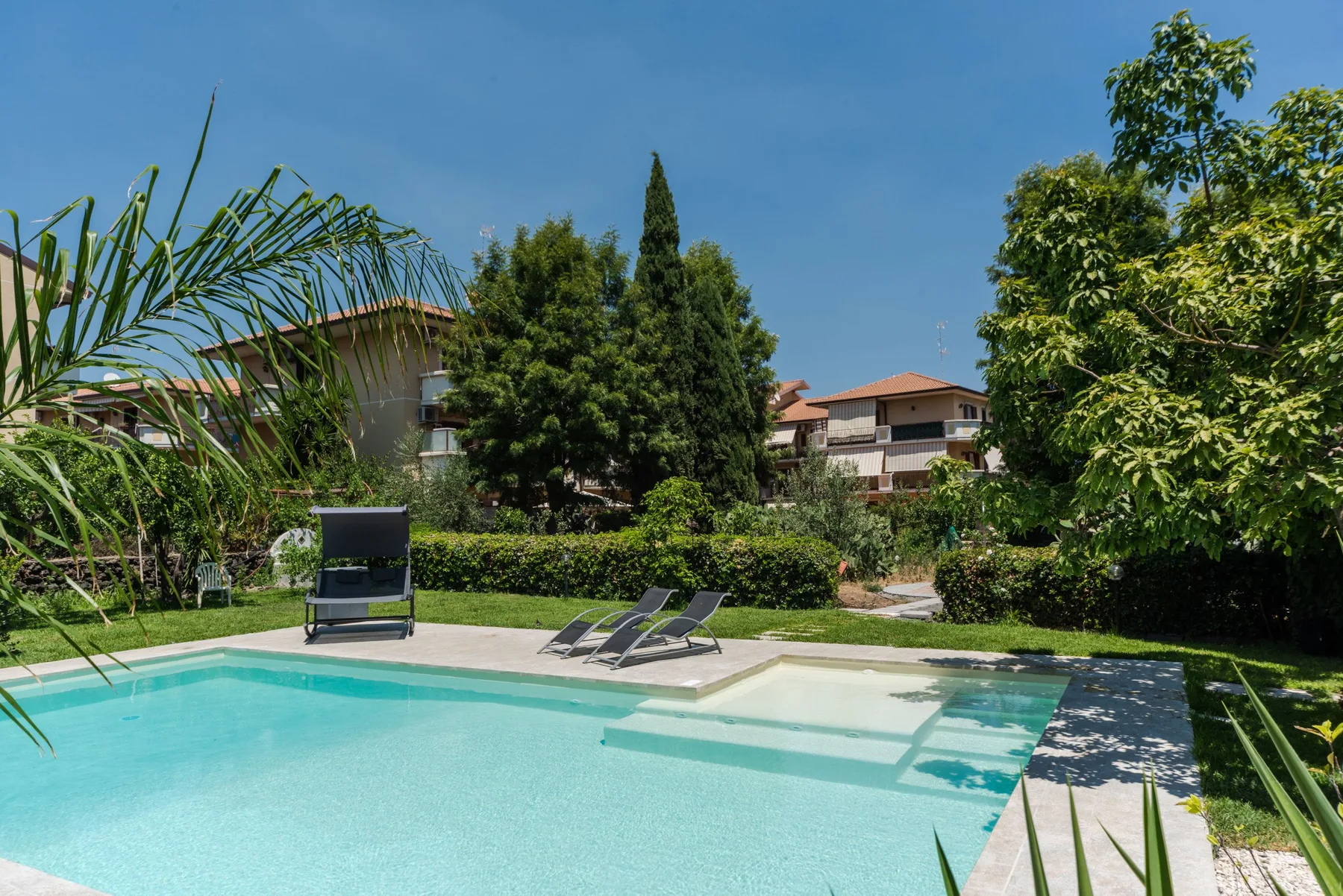 Una piscina al aire libre está rodeada de vegetación y edificios residenciales.