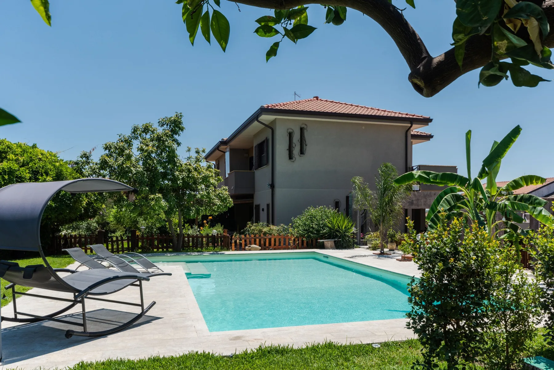 Una piscina en el patio está junto a una casa, con asientos y vegetación alrededor.