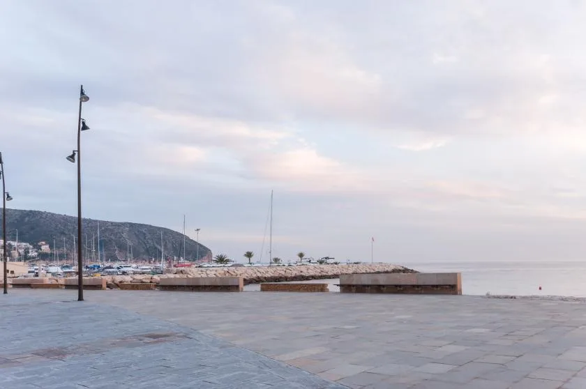 A paved waterfront promenade overlooks a marina and calm sea beneath a cloudy sky.