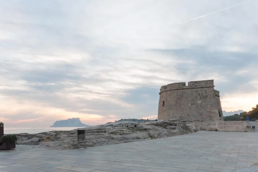 A stone coastal tower stands beside a paved waterfront under a cloudy sky.