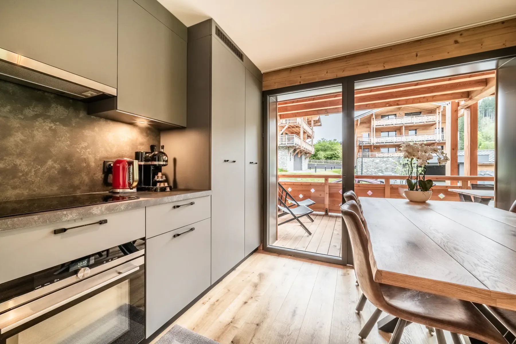 Cocina moderna con armarios grises, electrodomésticos de acero inoxidable y una mesa de comedor de madera. Unas grandes puertas correderas de cristal dan a un patio con muebles de madera y vistas a los edificios vecinos.