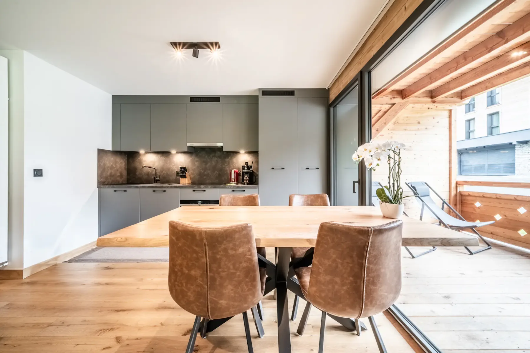 Un comedor con una mesa de madera y asientos tapizados en marrón junto a grandes ventanales.