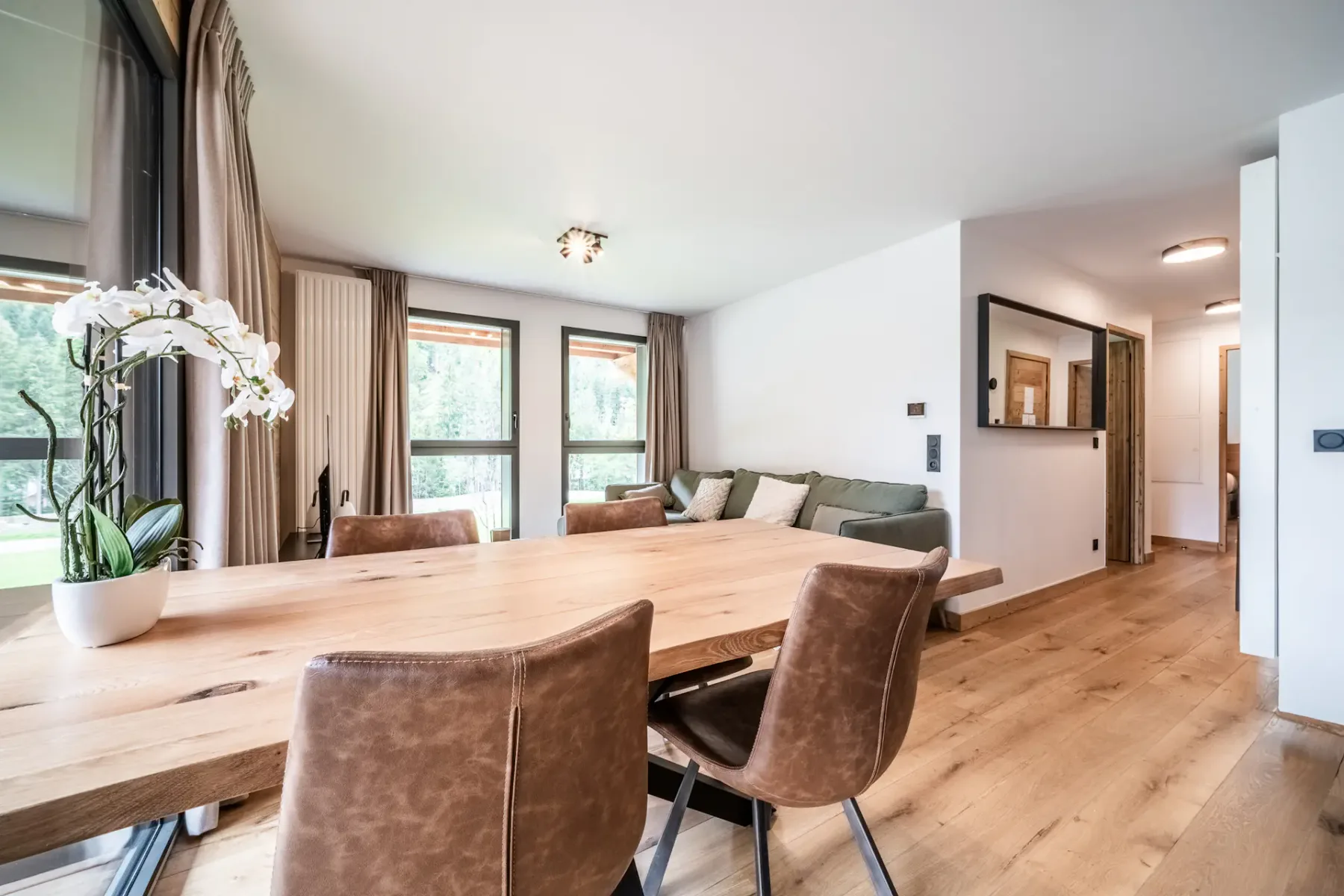 Una zona de comedor moderna con una mesa de madera y sillas de cuero marrón. Al fondo hay un sofá blanco cerca de grandes ventanas con cortinas beige. La estancia tiene suelo de madera y paredes claras.