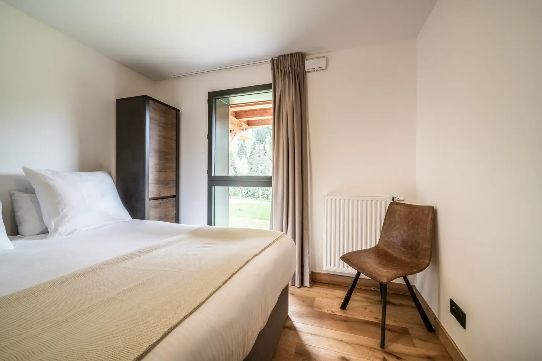 Un dormitorio con una cama, un armario, una ventana con cortinas y una silla junto a un radiador.