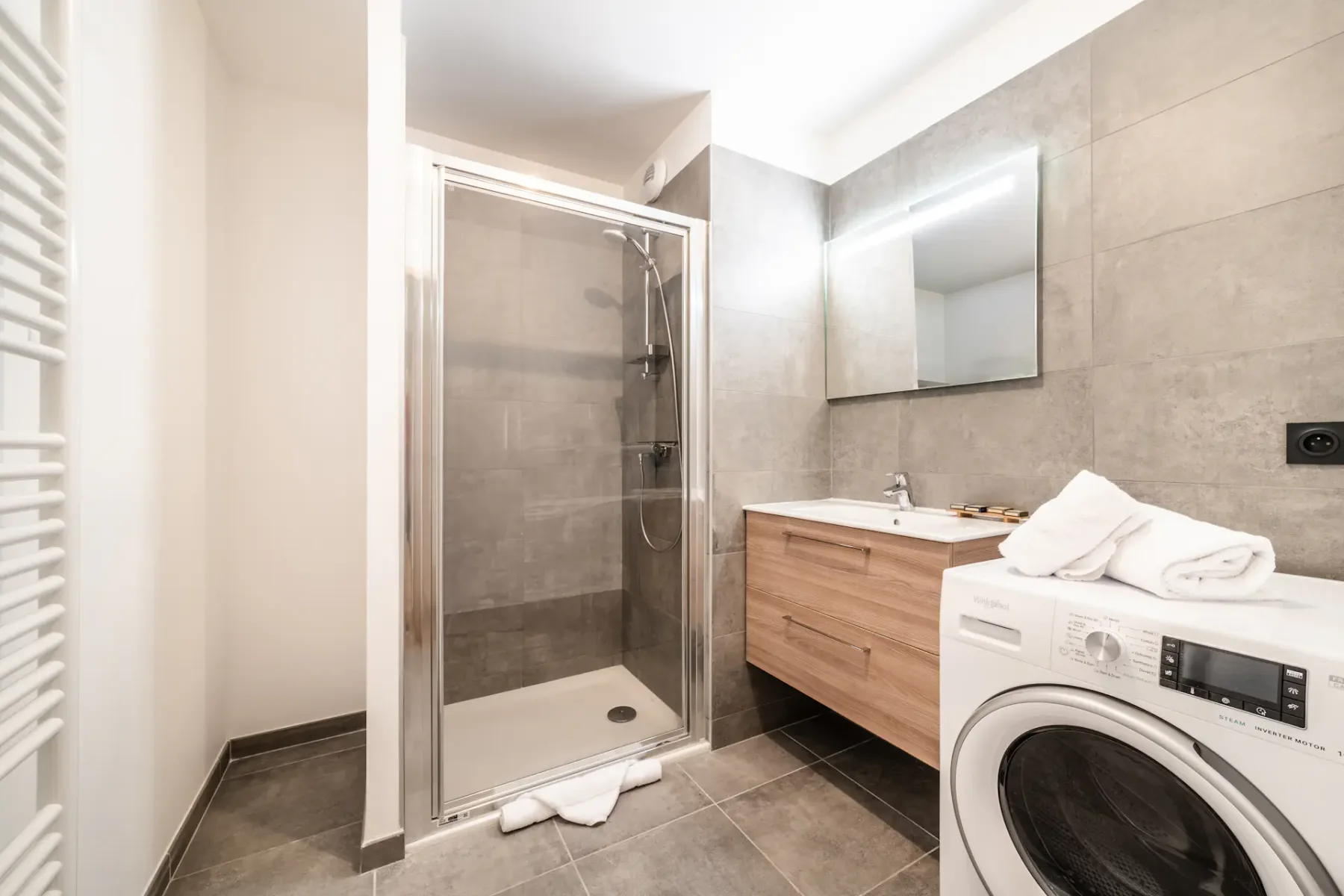 Baño moderno con una ducha con mampara de cristal, un mueble de lavabo de madera y una lavadora. Las paredes y el suelo están revestidos con baldosas grises. Hay un espejo sobre el lavabo y toallas dobladas sobre la lavadora.