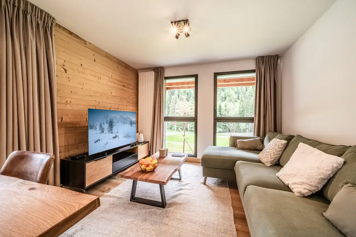 A living room with a television, coffee table, and seating beside tall windows.