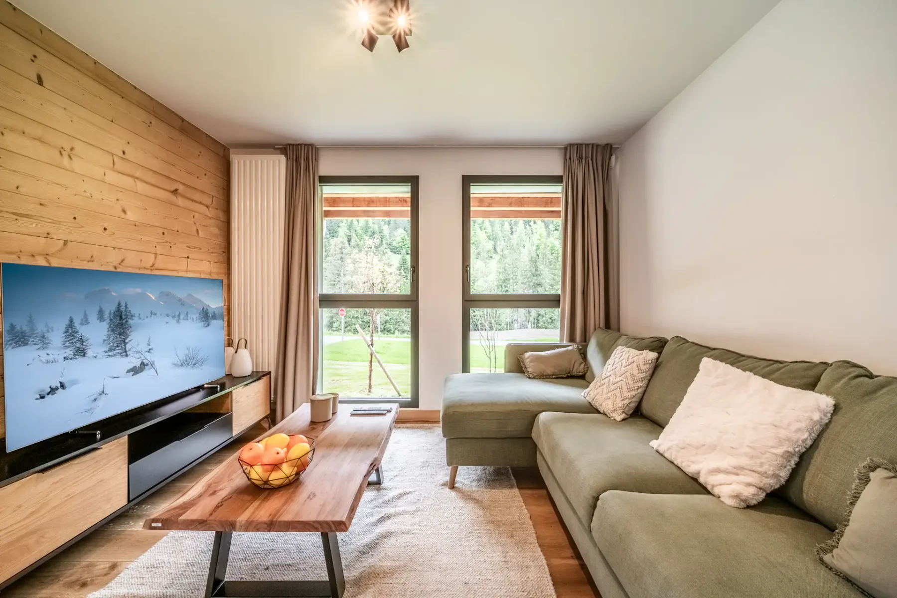 A cozy living room featuring a green sectional sofa, a wooden coffee table, a TV on a stand, and two large windows with a view of a green outdoor area. The decor includes brown curtains and pillows.