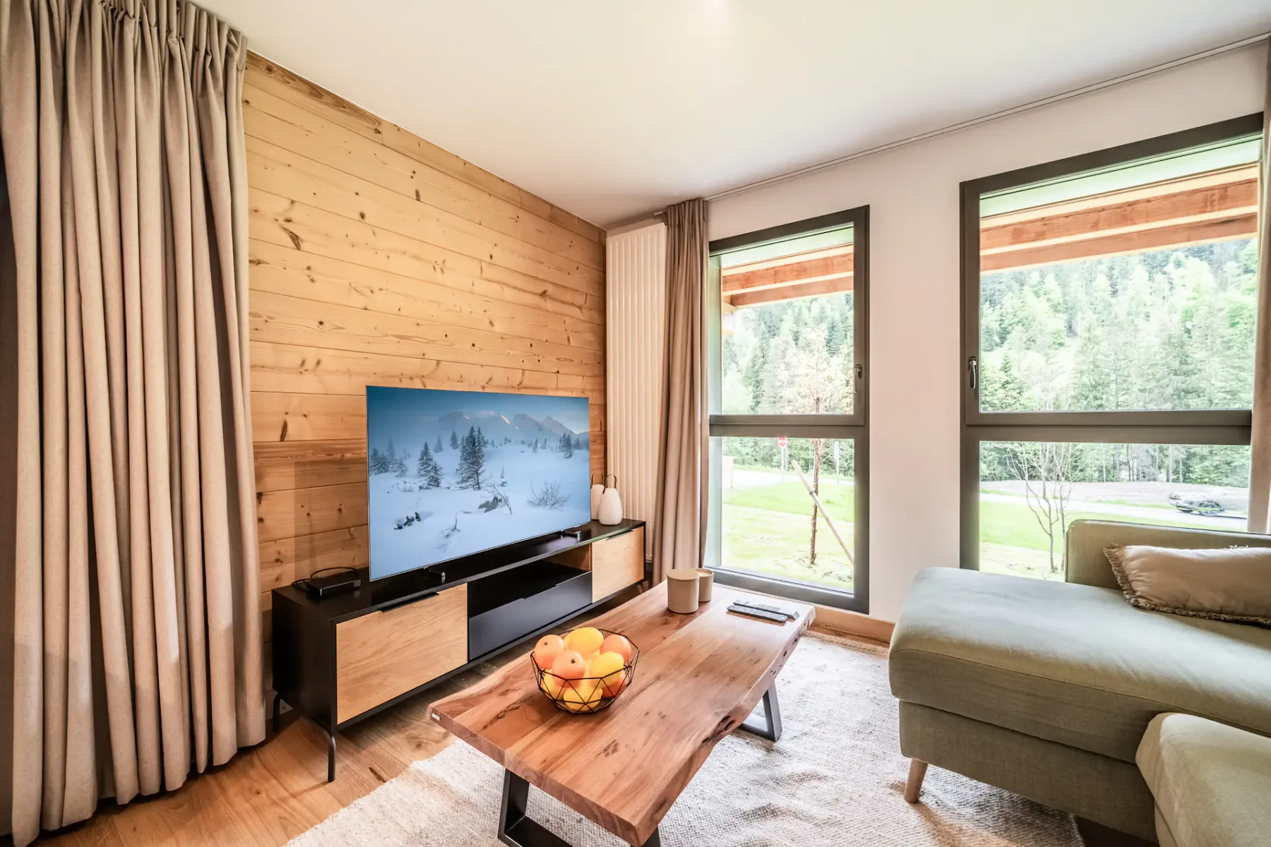 A living room with a television, wooden coffee table, and large windows overlooking trees.