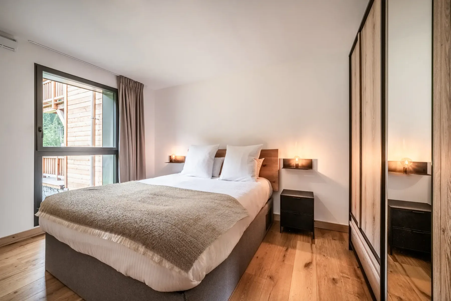 Un dormitorio bien ordenado con una cama grande, suelo de madera, mesas de noche, un armario con espejos y una ventana que aporta luz natural.