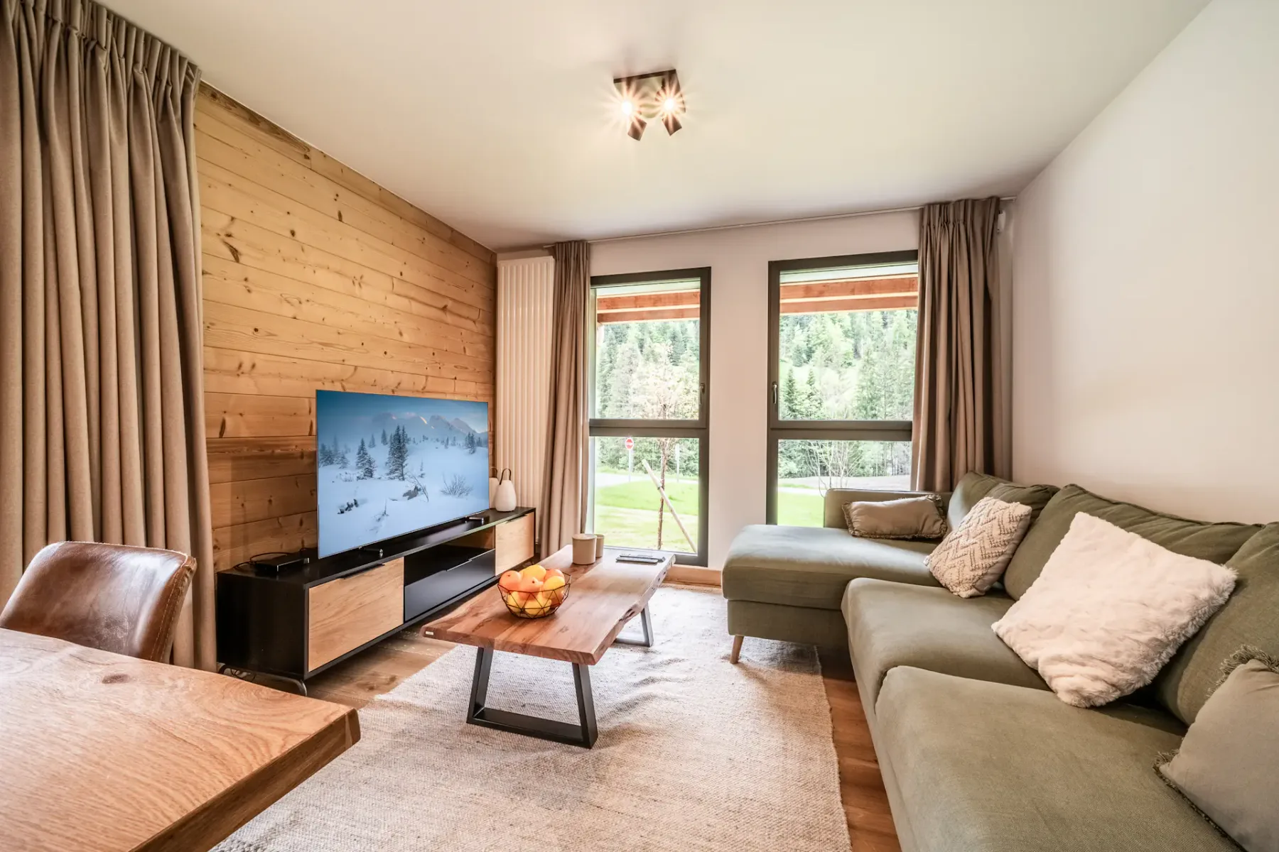 Una acogedora sala de estar con un sofá modular verde, una mesa de centro de madera, un televisor sobre un mueble y dos grandes ventanas con vistas a una zona verde exterior. La decoración incluye cortinas y cojines marrones.