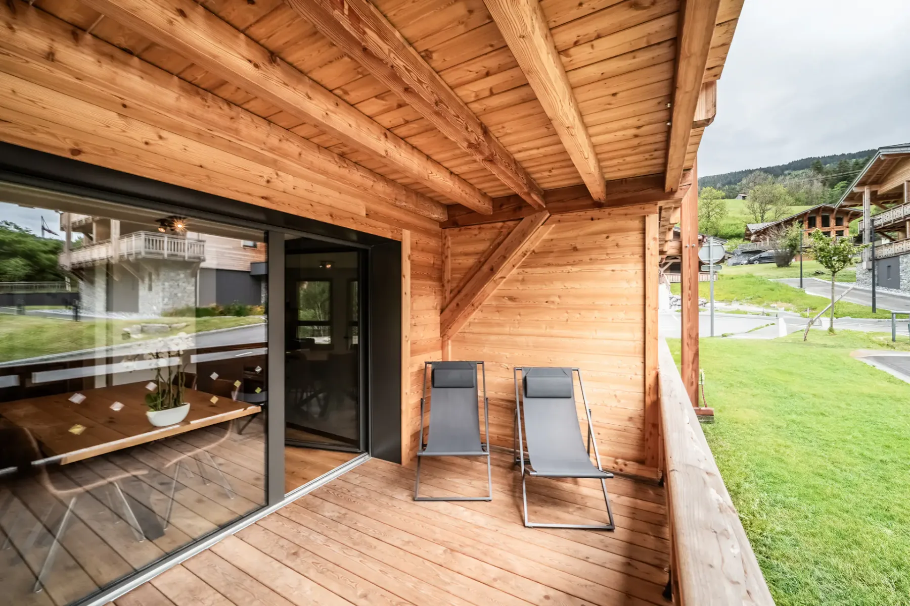 Una terraza de madera con dos tumbonas negras junto a una puerta corredera de cristal por la que entra luz. La terraza tiene vistas a una zona de césped y al fondo se ven casas vecinas.