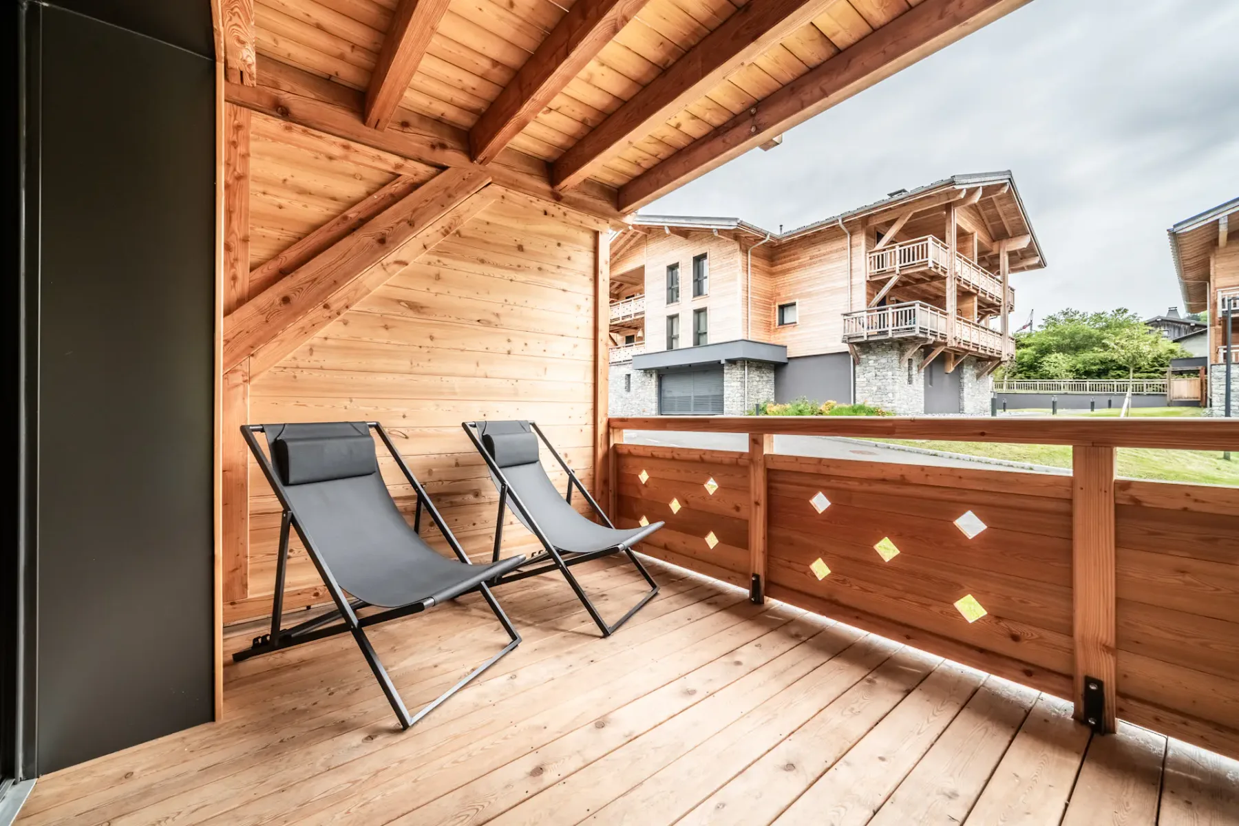 Una terraza cubierta de madera tiene dos sillas reclinables y una barandilla con edificios cercanos al fondo.