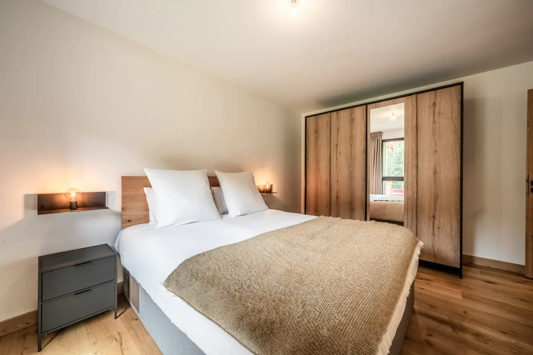 Un dormitorio bien ordenado con una cama grande, suelo de madera, mesas de noche, un armario con espejos y una ventana que aporta luz natural.